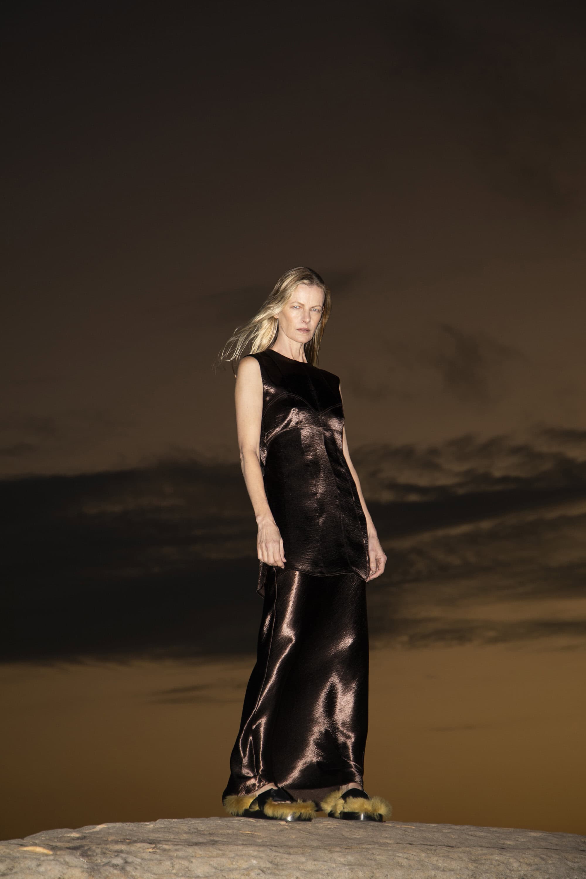 A woman poses outdoors in a dark, silky sleeveless top and matching maxi skirt. The lighting casts shadows and reflects off the fabric.