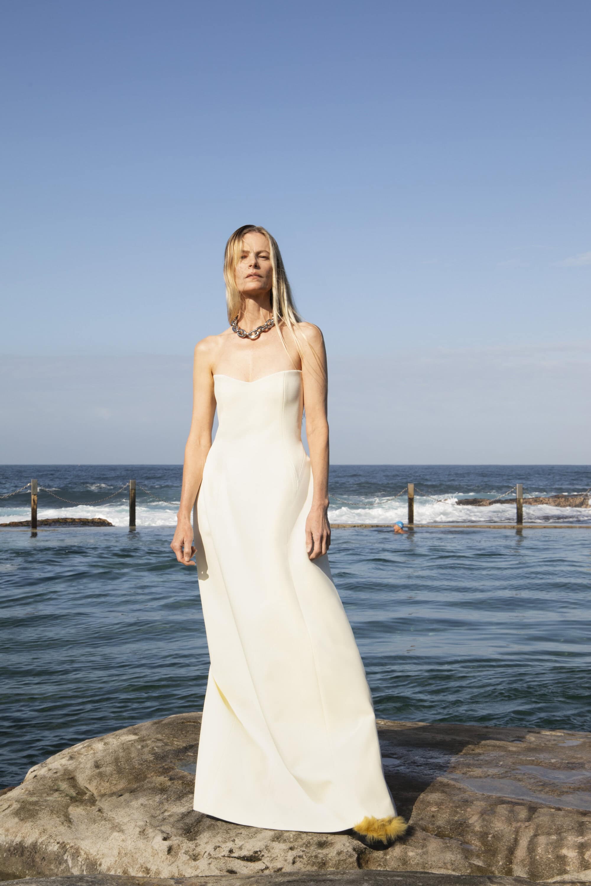 Elegant woman in a white gown poses on rocks by the ocean under a clear sky, accessorized with a chunky silver necklace and feather trim detail.