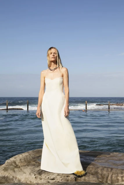 Elegant woman in a white gown poses on rocks by the ocean under a clear sky, accessorized with a chunky silver necklace and feather trim detail.