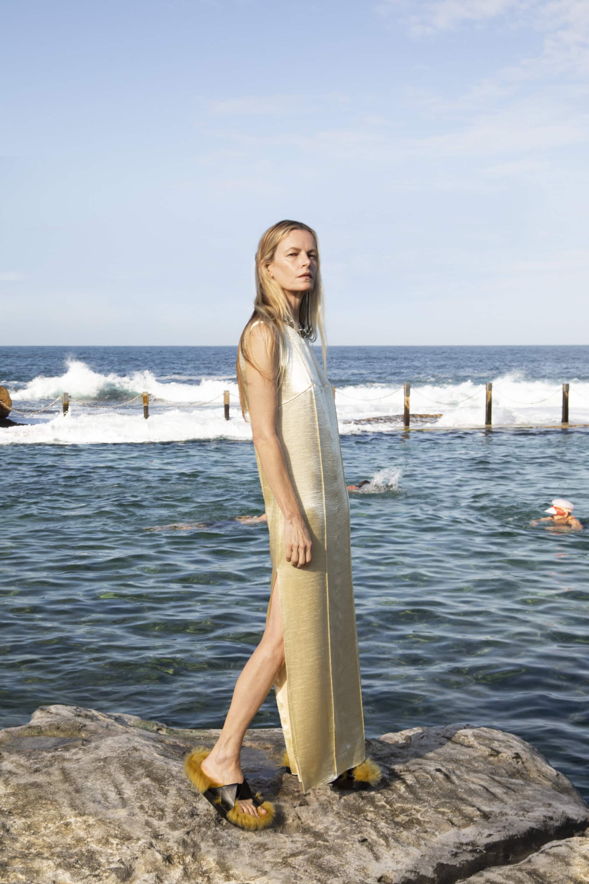 Woman in a long, light gold dress and platform sandals poses on rocks near the ocean on a sunny day. Sky and swimmers visible.