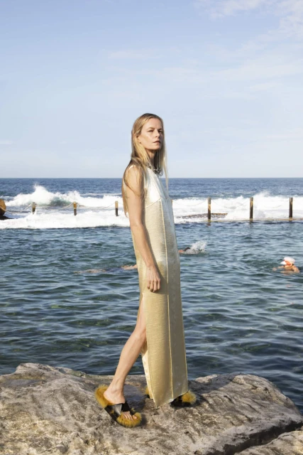 Woman in a long, light gold dress and platform sandals poses on rocks near the ocean on a sunny day. Sky and swimmers visible.