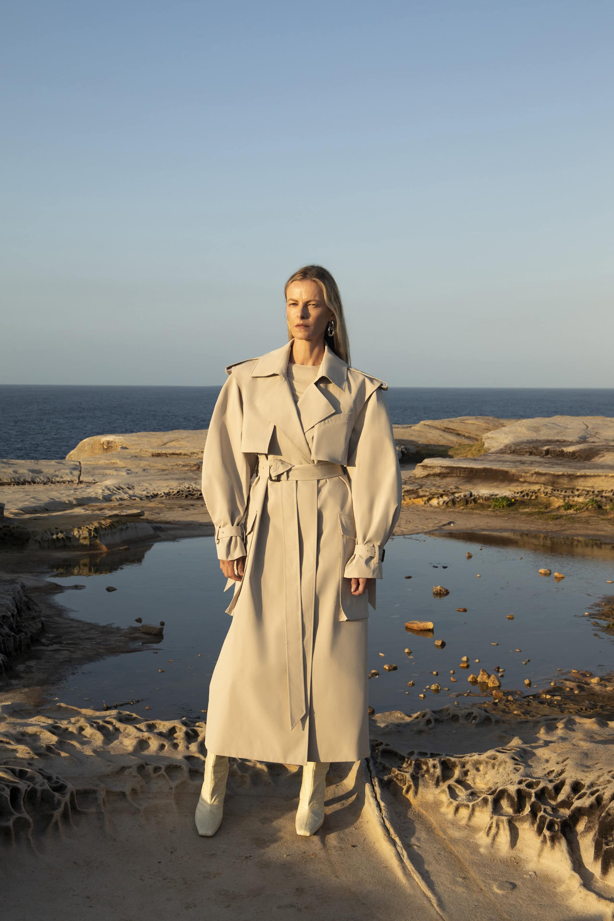 Elegant woman in a trench coat and heeled boots poses on a rocky coastline with the sea and sky in the background on a sunny day.