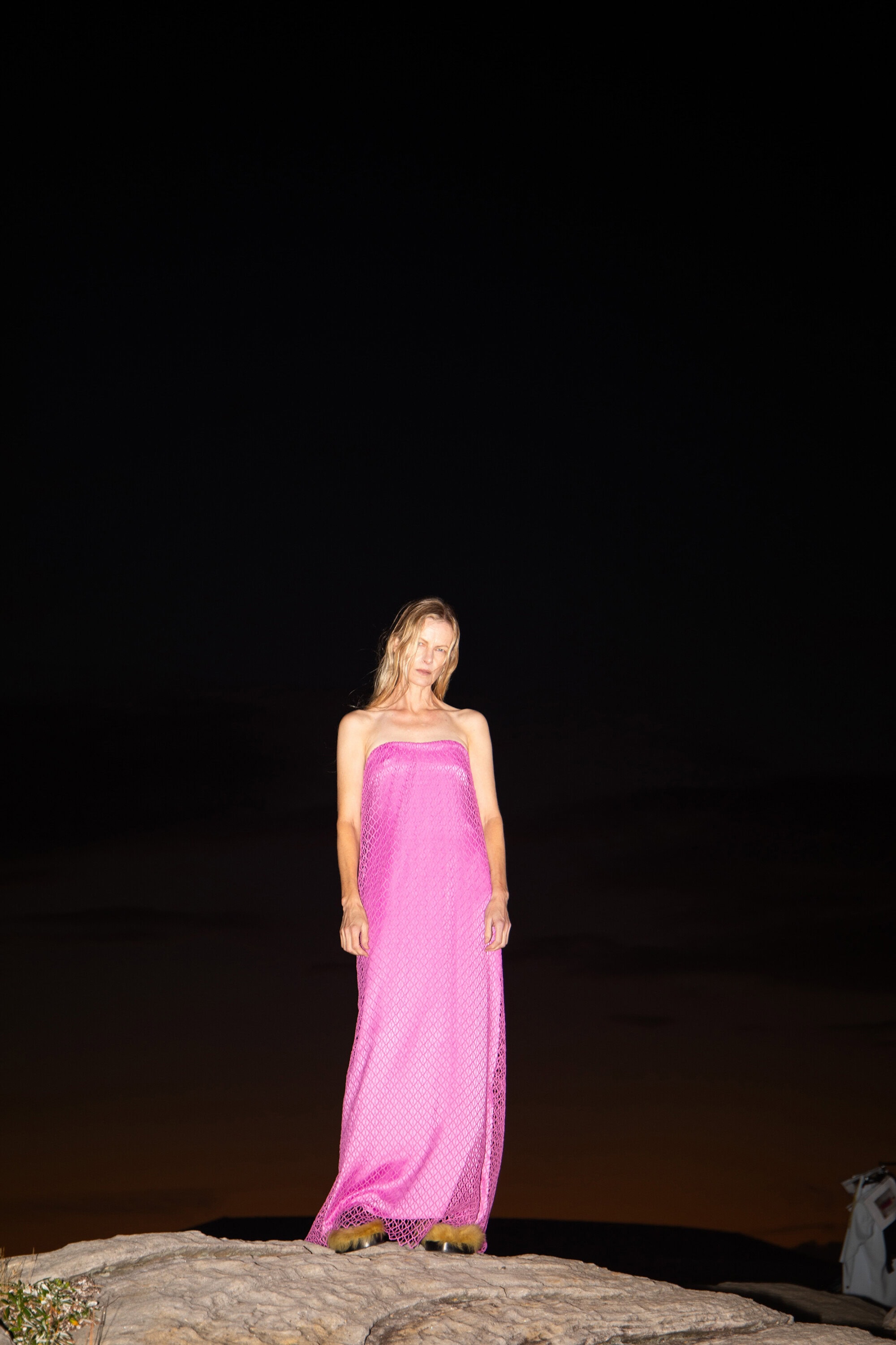 Blonde woman in pink dress stands on a rock at night. The shot is a full-length portrait with a dark sky background.