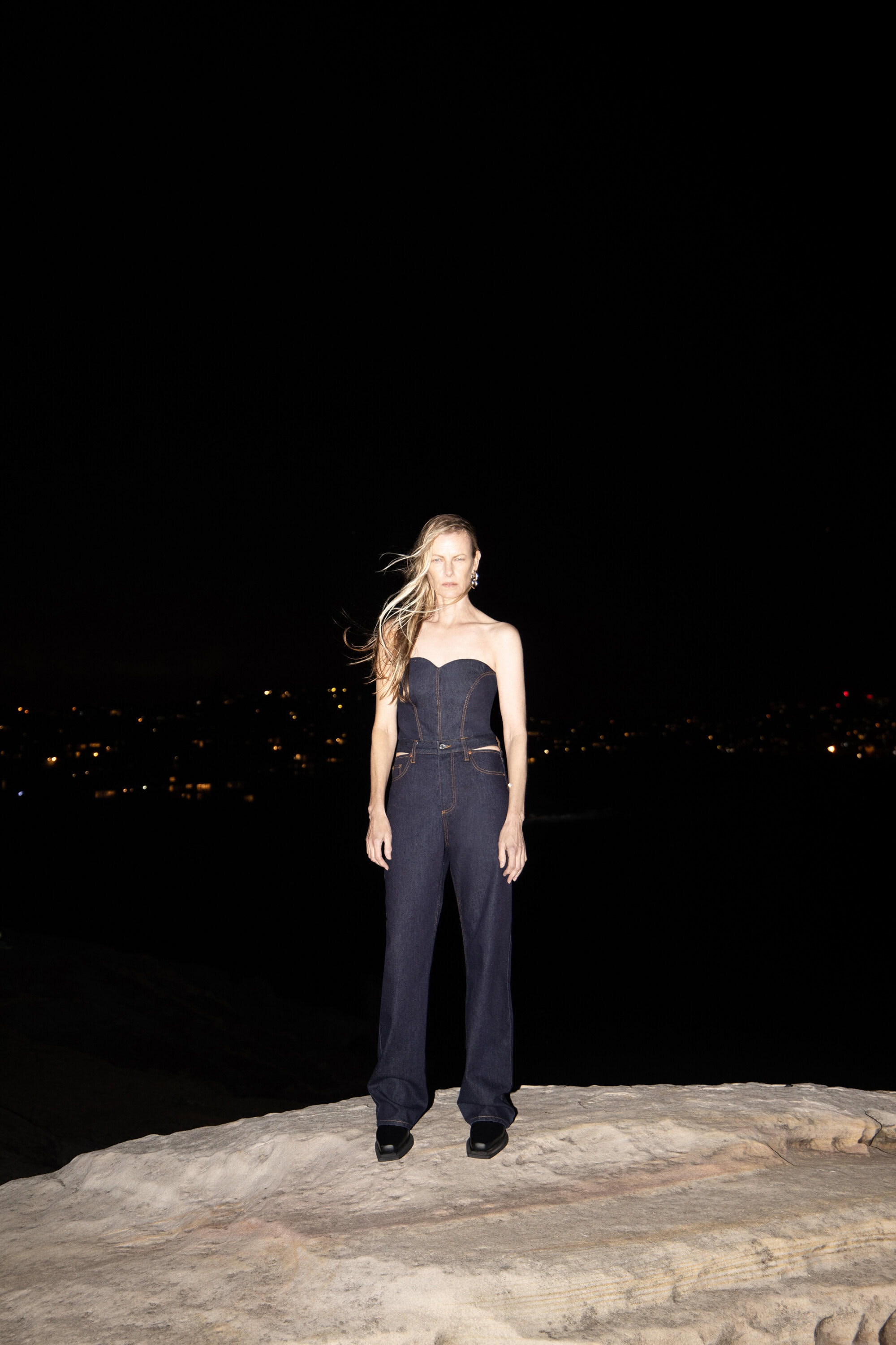 Model wearing dark denim strapless top and matching trousers standing on a rocky outcrop at night.