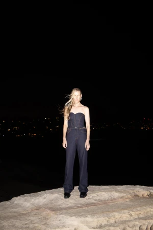 Model wearing dark denim strapless top and matching trousers standing on a rocky outcrop at night.