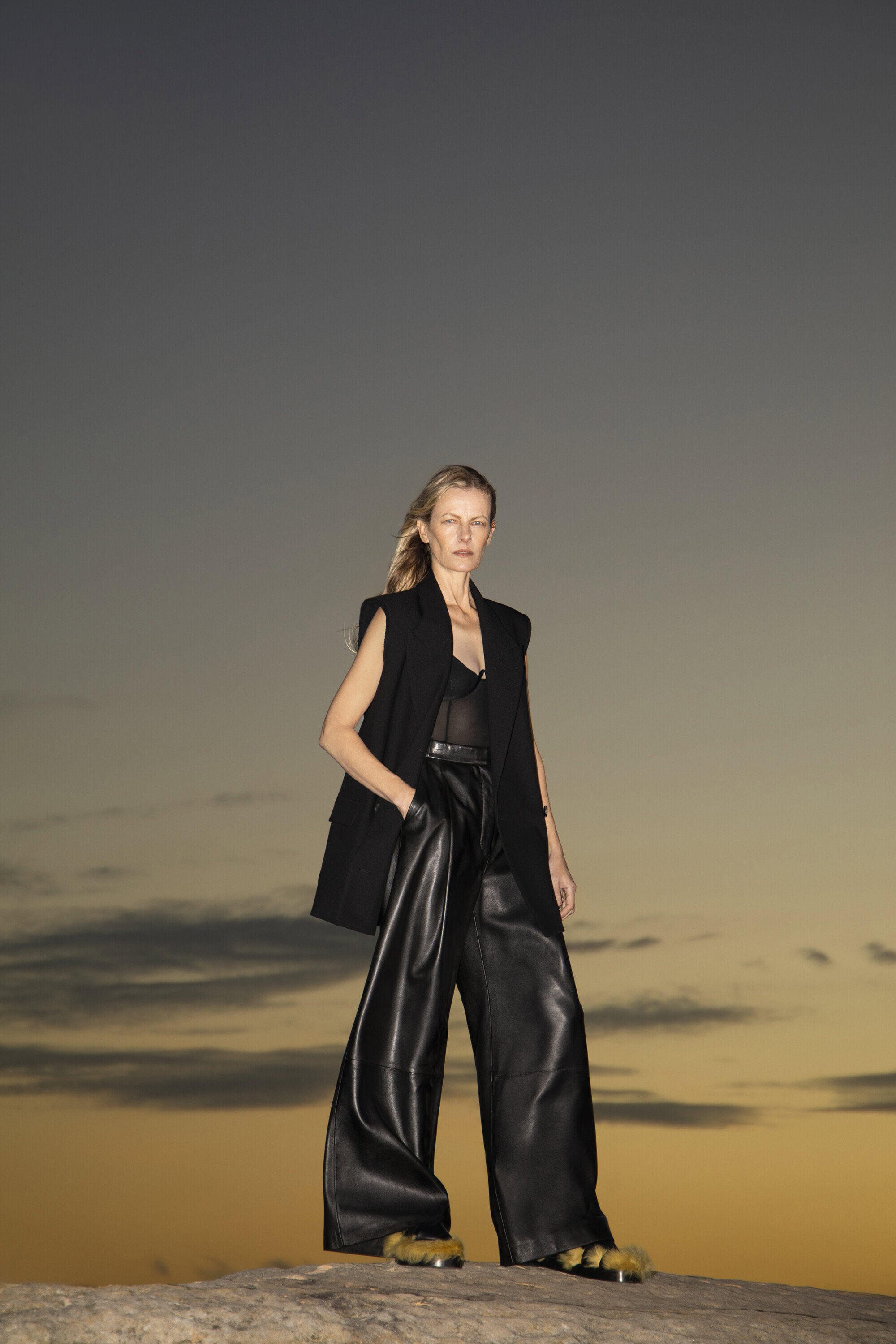 A woman poses in a black vest and wide-leg leather pants, standing on a rock formation against a soft sunset sky.
