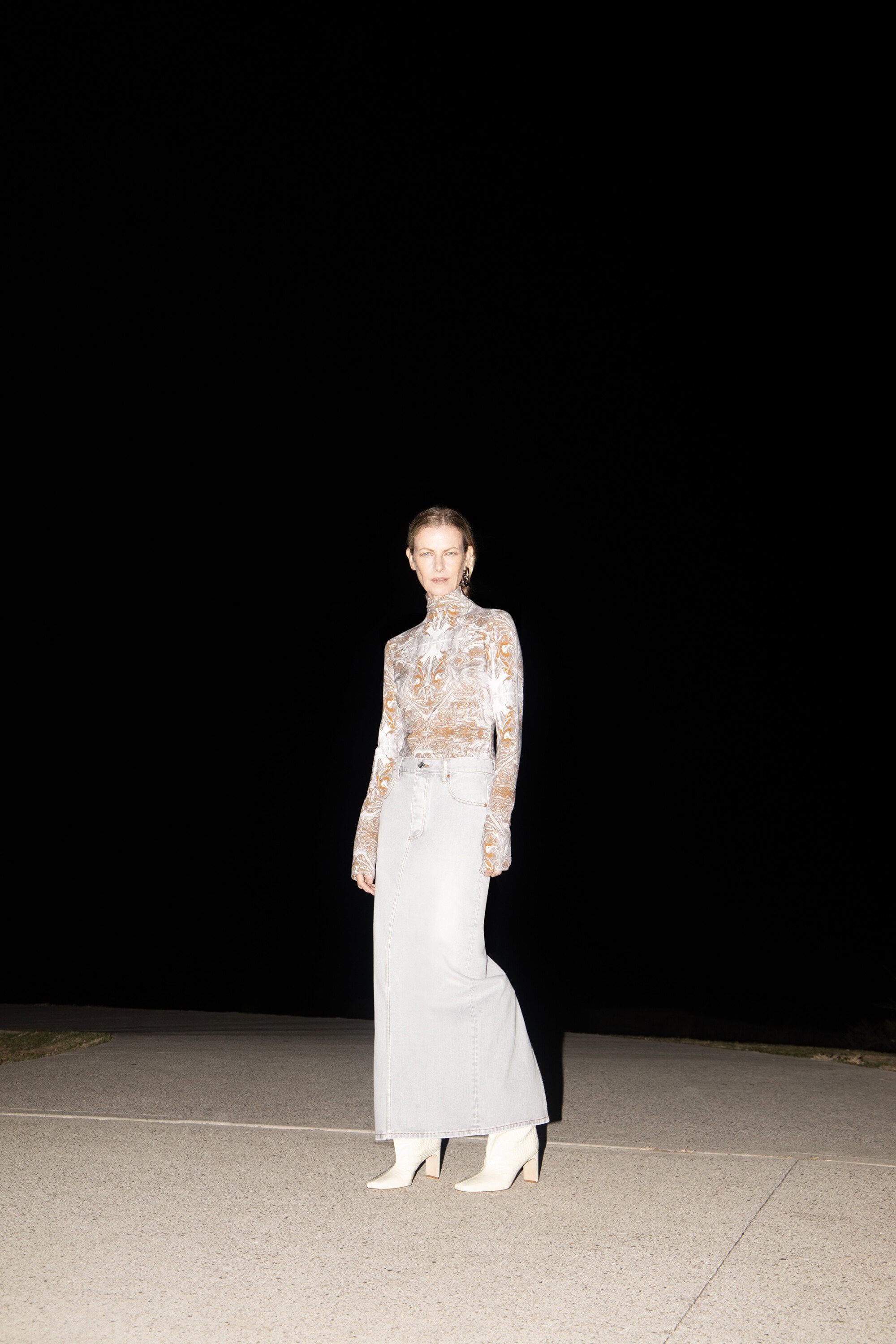 Adult female fashion model poses outdoors in a long skirt, boots, and lace top against a dark background in a medium shot.