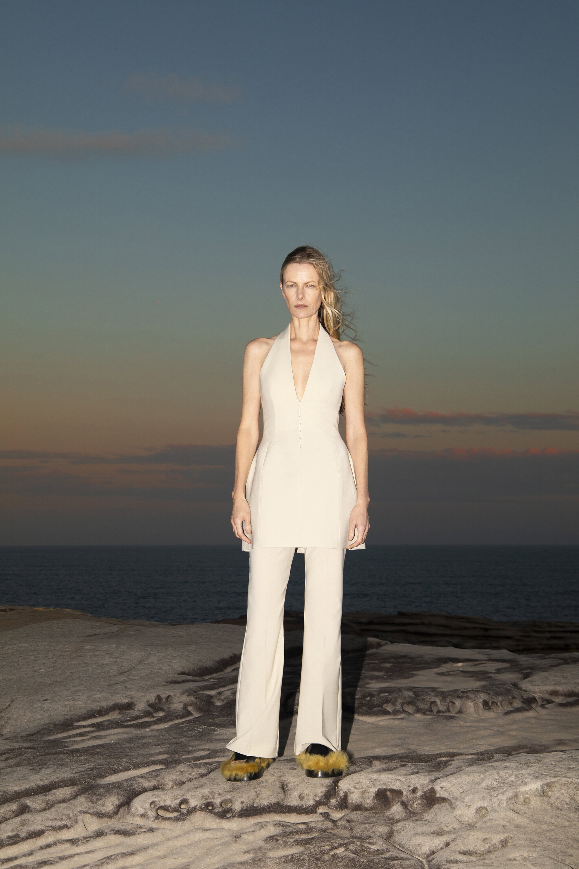 Woman in white halter top and pants poses on rocky coast at sunset with ocean backdrop. Fashion photography in natural setting.