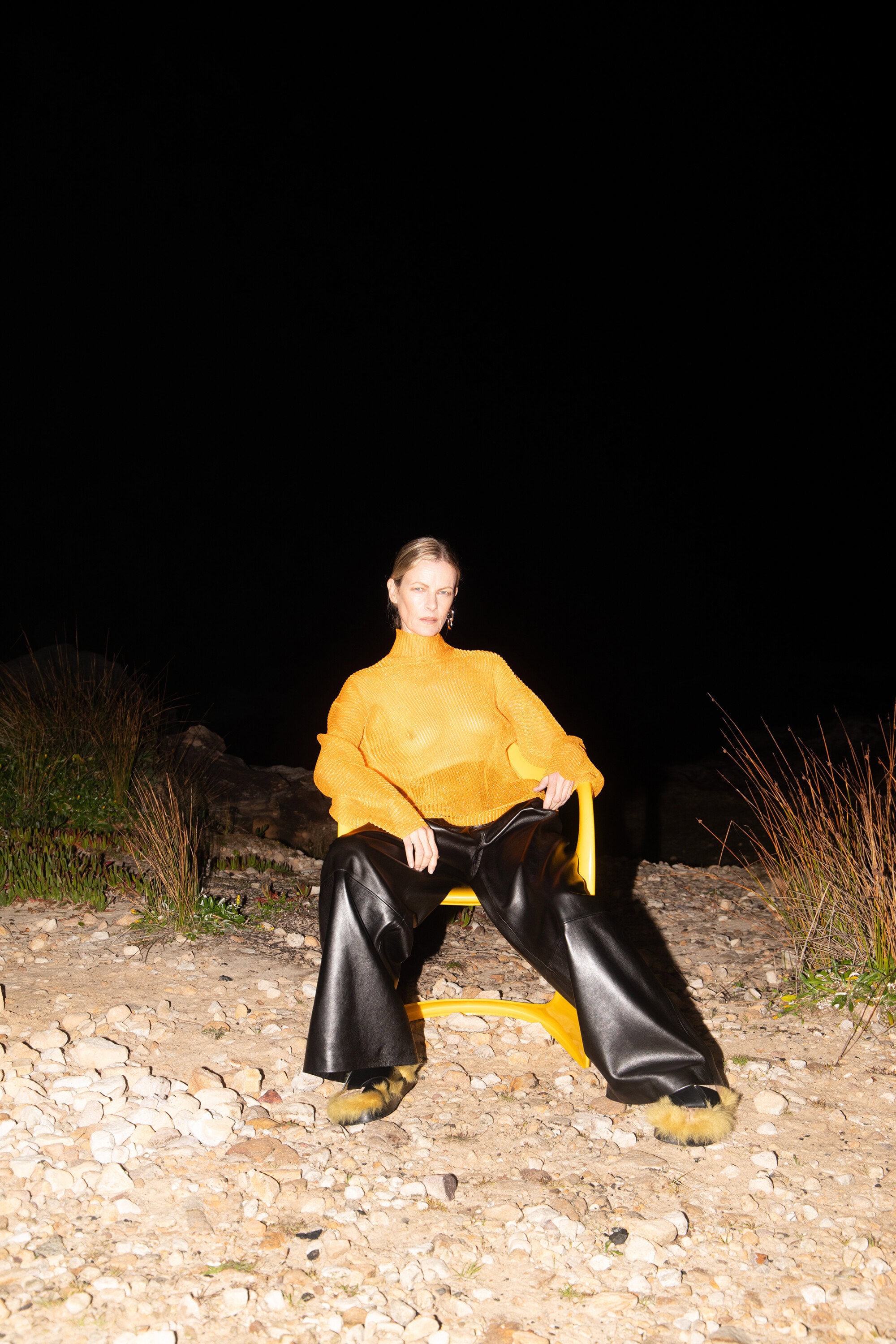 Fashion photo of a woman in an orange turtleneck and black pants sitting on a yellow chair on a rocky surface outdoors at night.