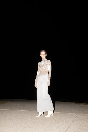 Model Emma Balfour standing at night in a patterned turtleneck top, light denim maxi skirt, and white heeled boots.