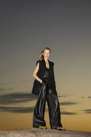 Model standing outdoors on rock wearing black sleeveless vest and leather trousers against overcast sky