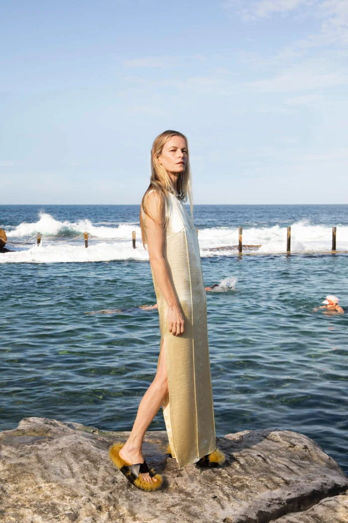 Emma Balfour standing on coastal rocks in a pale gold sleeveless maxi dress by an ocean pool at Maroubra Beach.