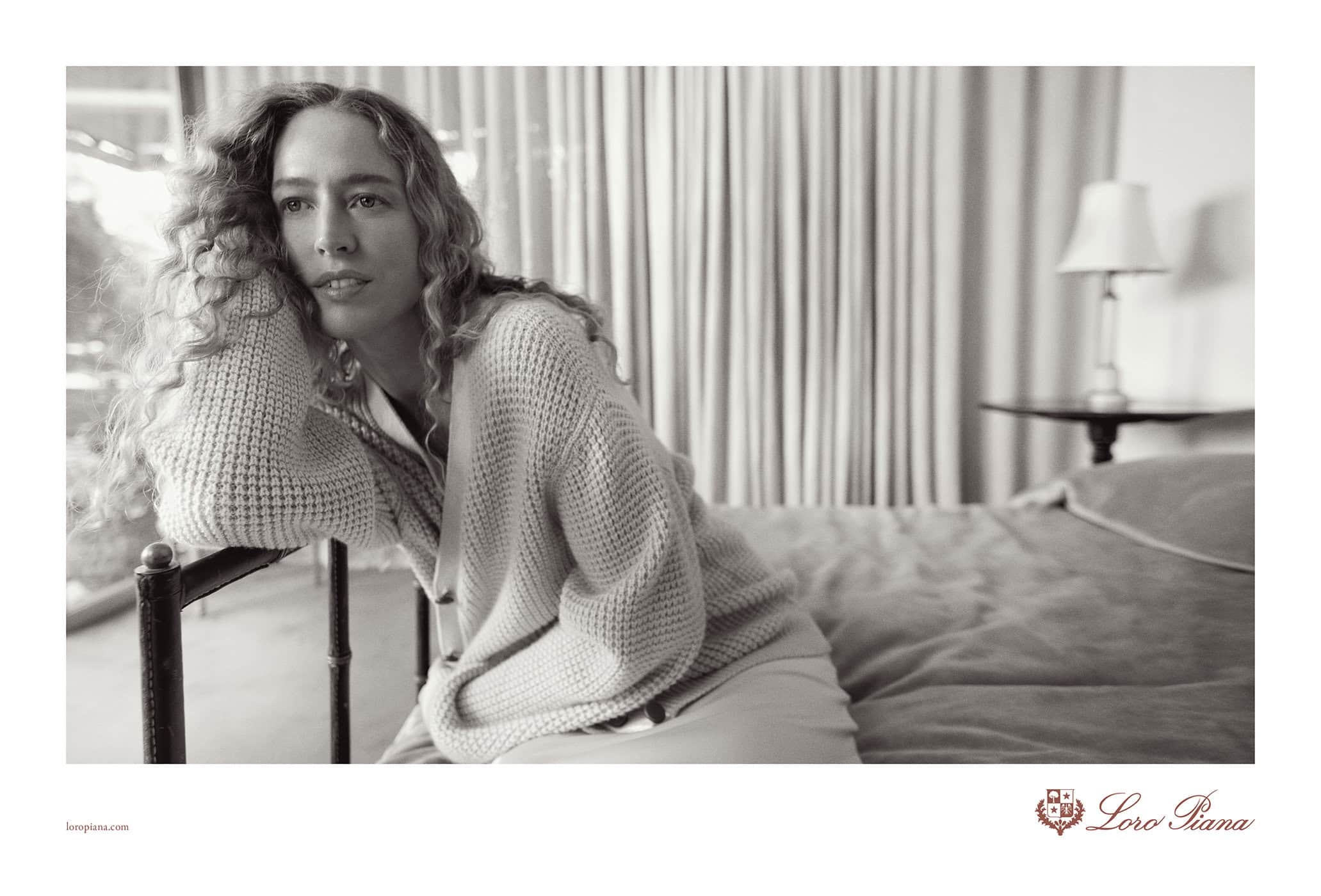 Monochrome portrait of a curly-haired woman in a cozy sweater, leaning on a bed frame in a softly lit indoor setting. Fashion photography.