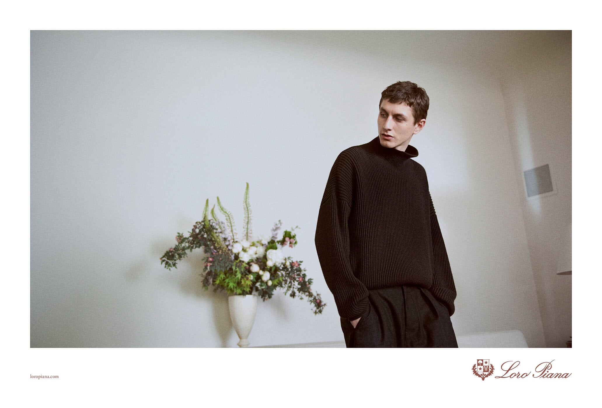 Fashion portrait of a male model in a black turtleneck sweater with flowers and Loro Piana logo on a white background in a photo studio.