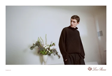 Man wearing a dark ribbed turtleneck sweater standing indoors beside a large floral arrangement for Loro Piana