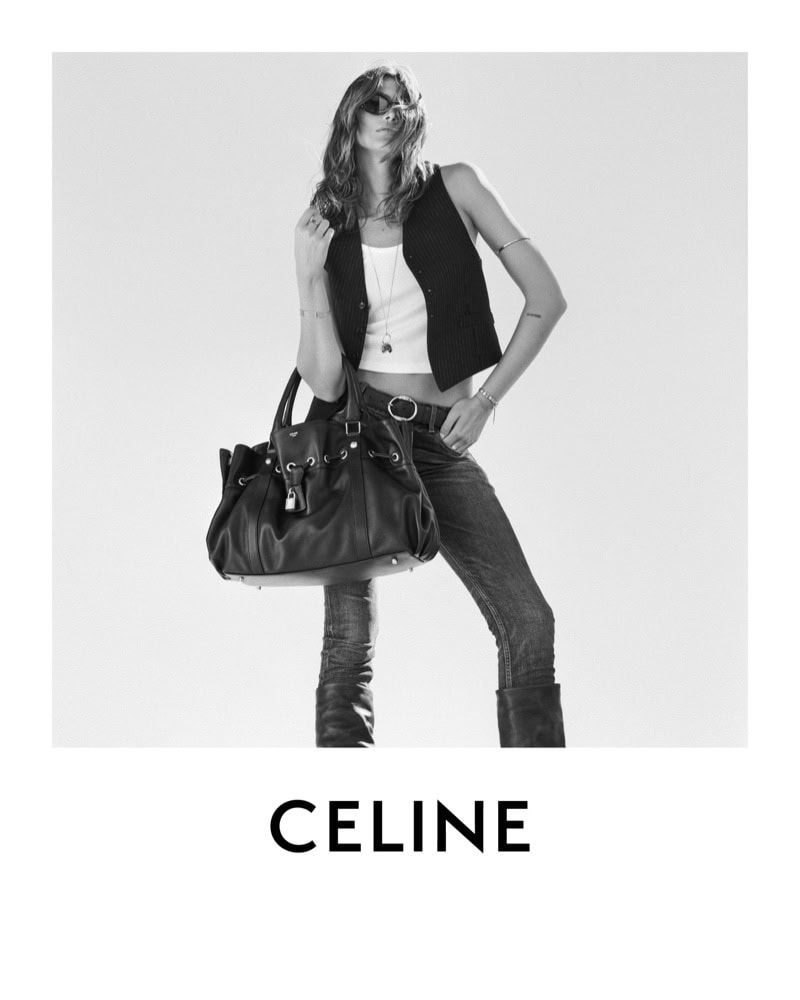 Black and white studio shot of a fashion model posing with a black leather purse against a bright background, wearing a vest, and jeans.