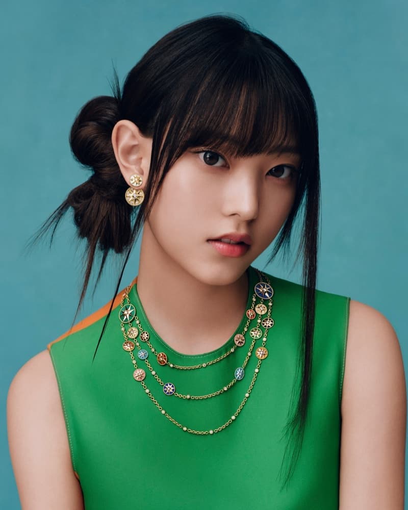 A young Asian woman poses in a sleeveless green top with layered gold necklace and matching earrings against a blue backdrop.