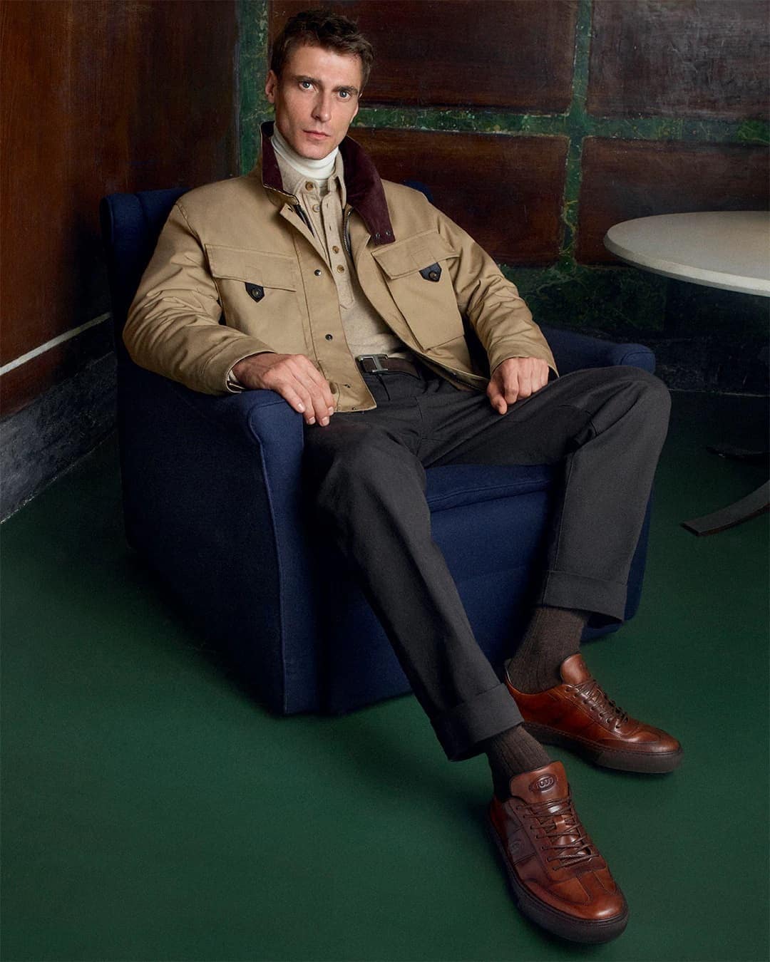 A well-dressed man in a jacket and trousers sits in a blue chair, complemented by his leather shoes and socks indoors with a dark green floor.