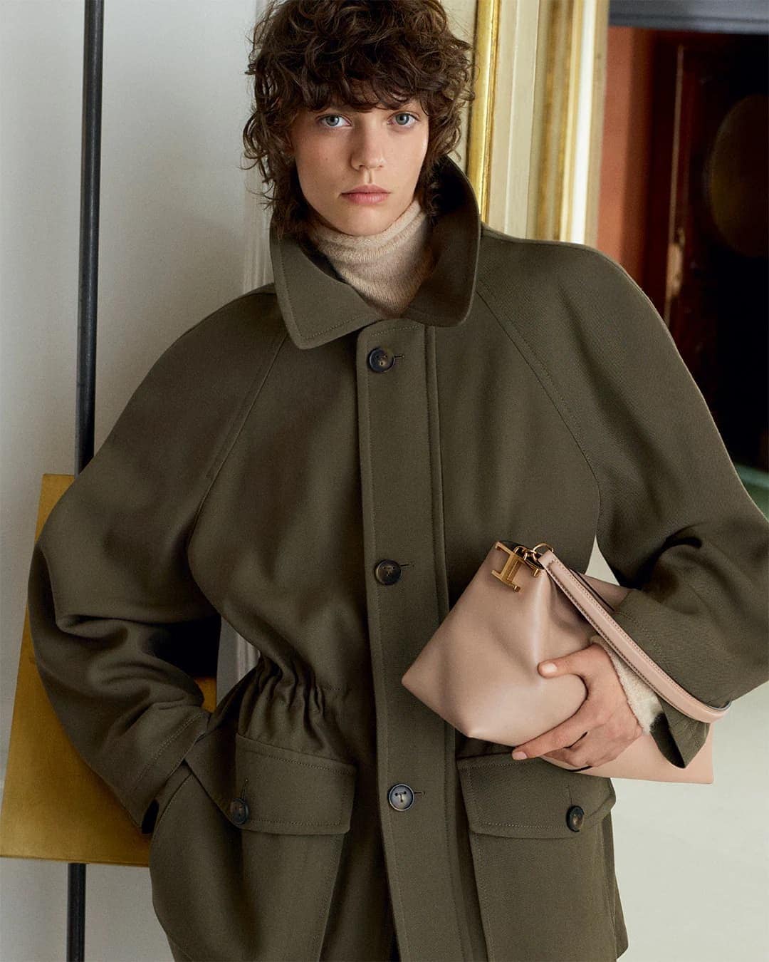 Fashion shot of a woman wearing an olive-green coat and holding a beige handbag, posing indoors with neutral background.