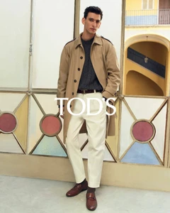 Model in camel trench coat and white trousers standing against a patterned architectural backdrop for Tod's ad campaign