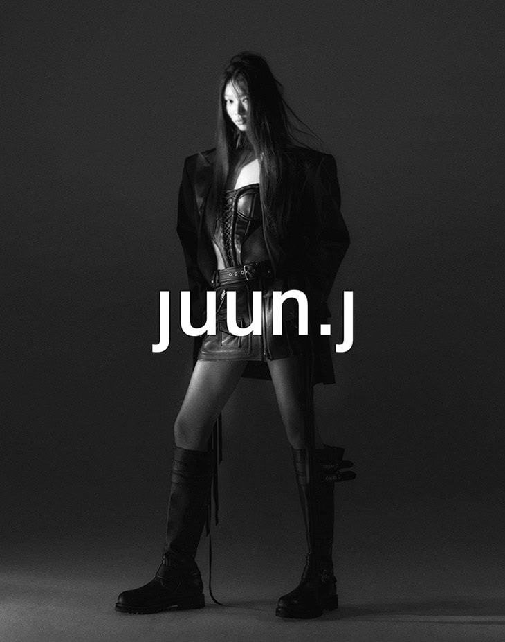 Black and white studio shot of a slender adult woman in a short leather skirt, long boots, and a blazer, with the text "juun.j".