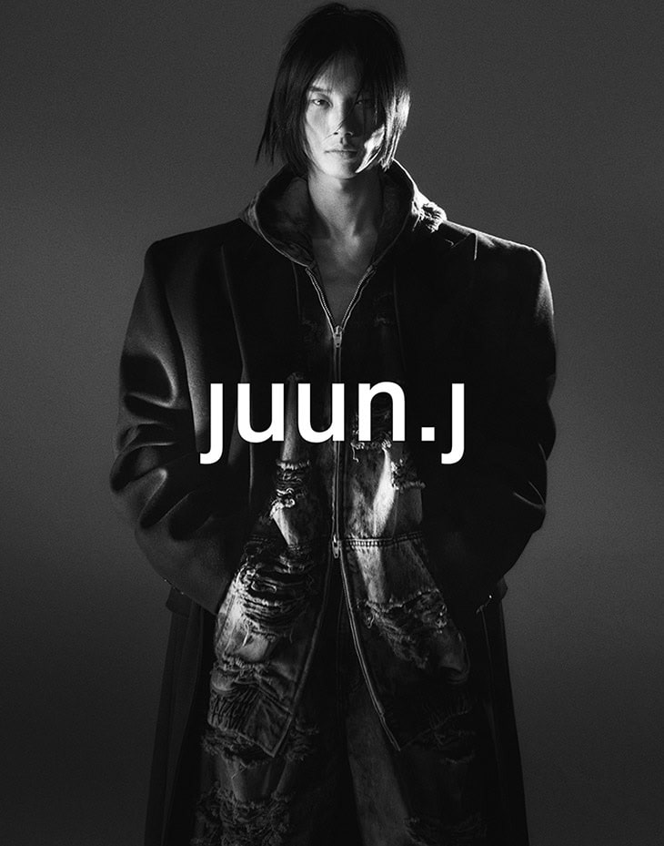 A person with dark mid-length hair wears an oversized black coat and distressed zip-up top in a low-key black and white portrait.