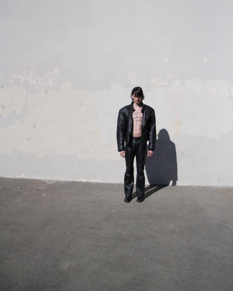 A slender adult man wearing a black leather jacket and pants stands in front of a weathered wall on a sunny day, casting a sharp shadow.