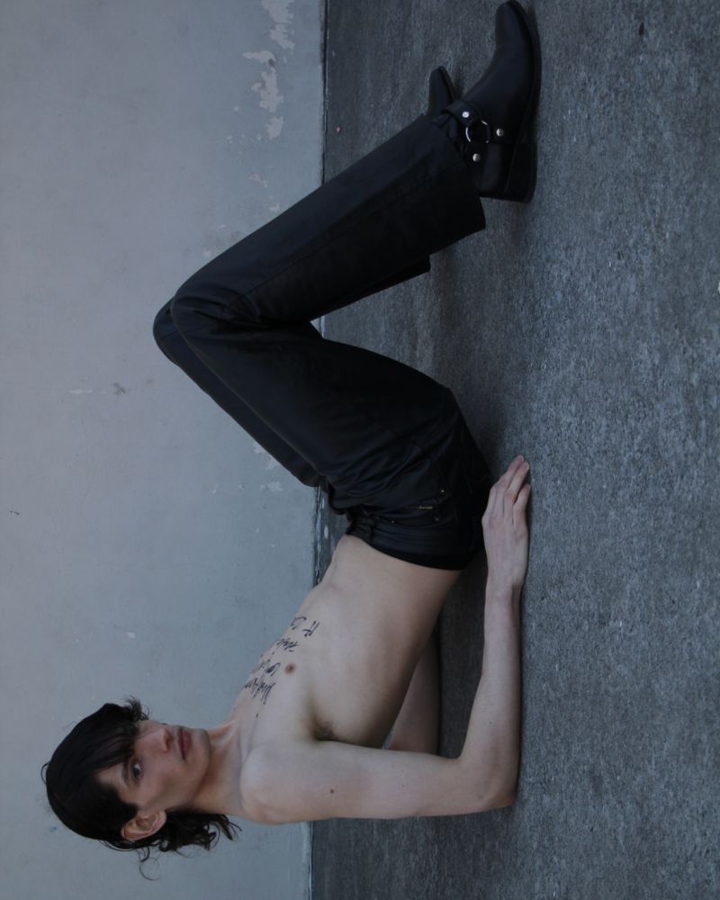 Young adult male model with black hair poses shirtless against a concrete wall, wearing dark pants and boots in a fashion editorial shoot.