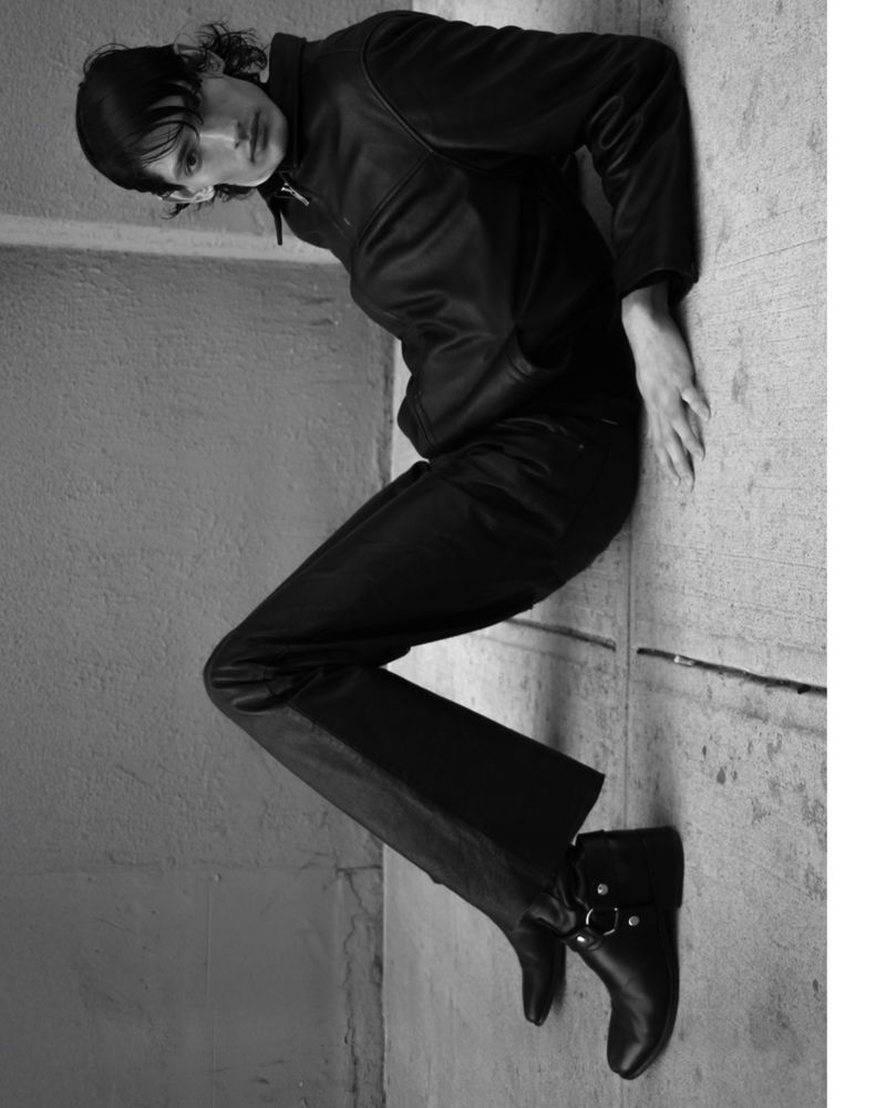 A person in dark clothing leans against a corner, legs extended, wearing boots, captured in a monochrome fashion shot with studio lighting.
