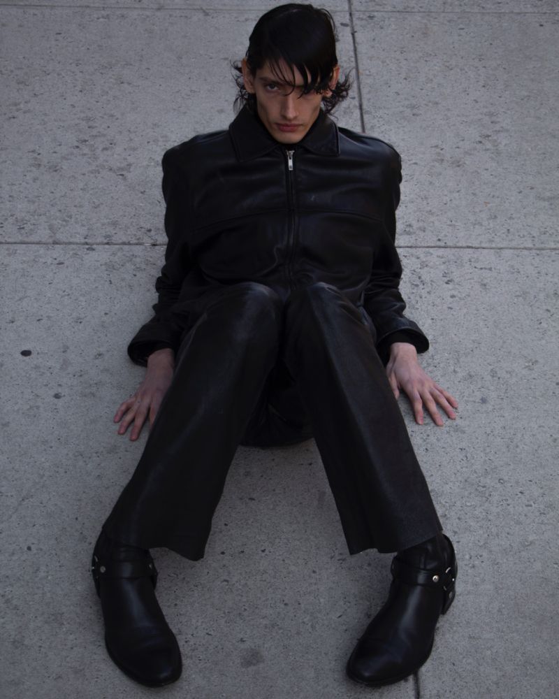 An androgynous adult poses outdoors on concrete in a black leather jacket, pants, and boots, illuminated by ambient light.