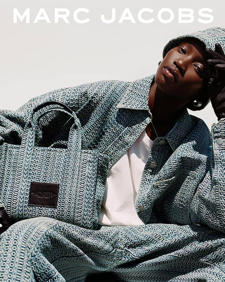 Fashion shot of a Black woman wearing a patterned jacket and bucket hat, holding a matching tote bag, posing against a white backdrop.