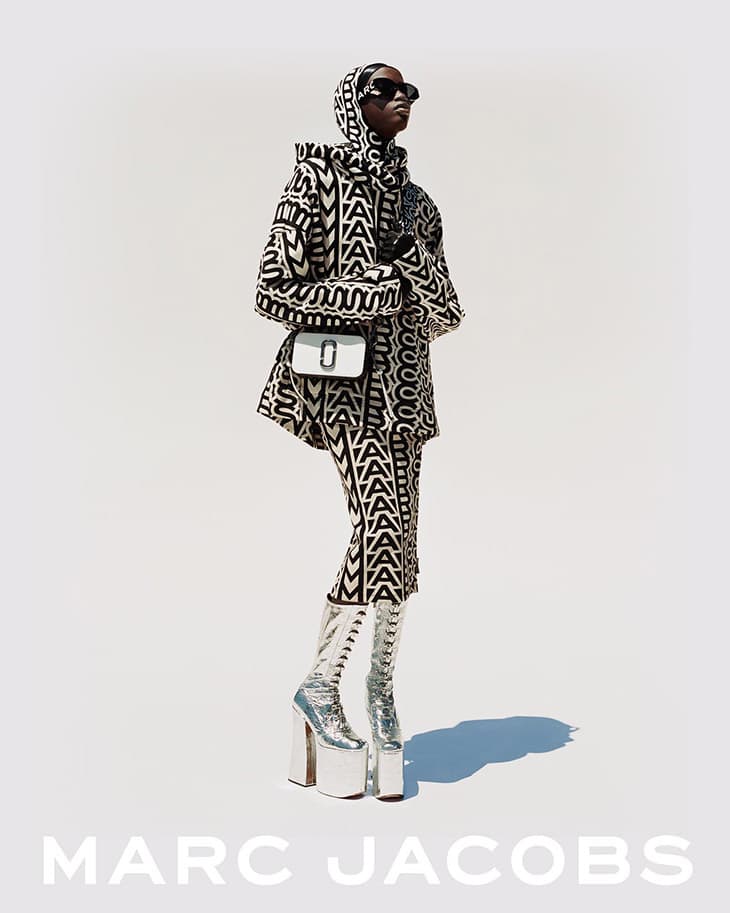 Model in patterned outfit, hood, sunglasses, and platform boots poses with a white purse against a white background for a fashion editorial.