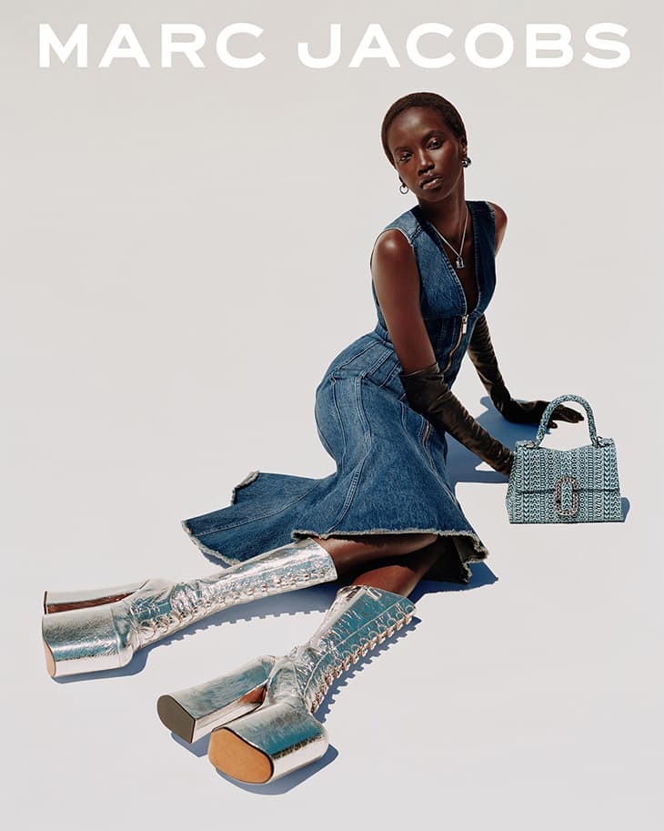 Fashion shot of an African-American woman wearing a denim dress and platform boots, posing with a patterned handbag against a white backdrop.