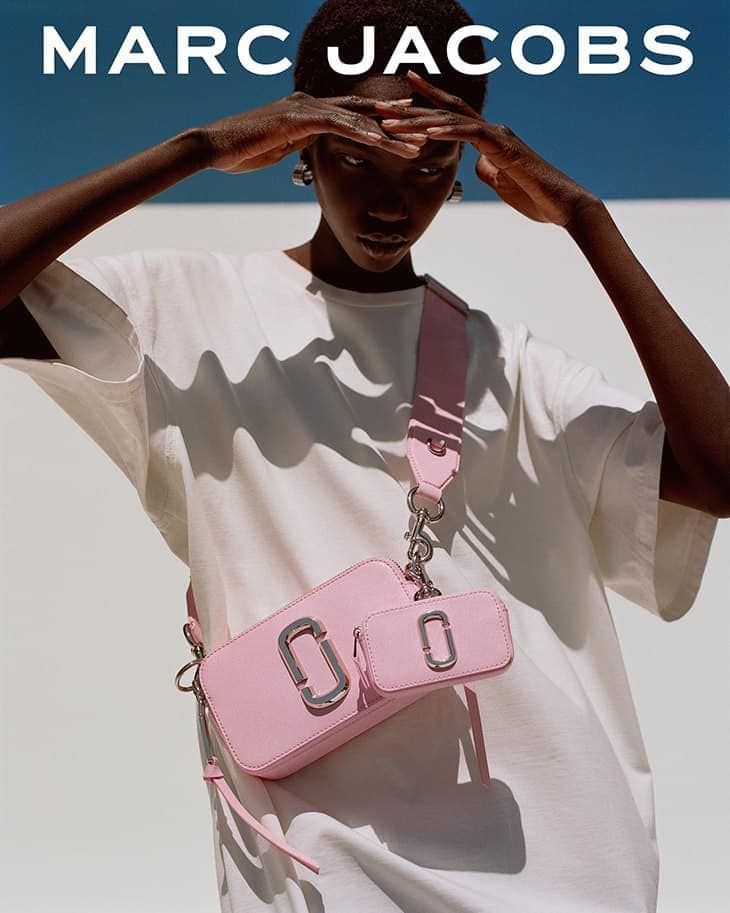 Fashion shot of a Black model wearing a white t-shirt and a pink purse with a metal logo against a blue sky and white wall.
