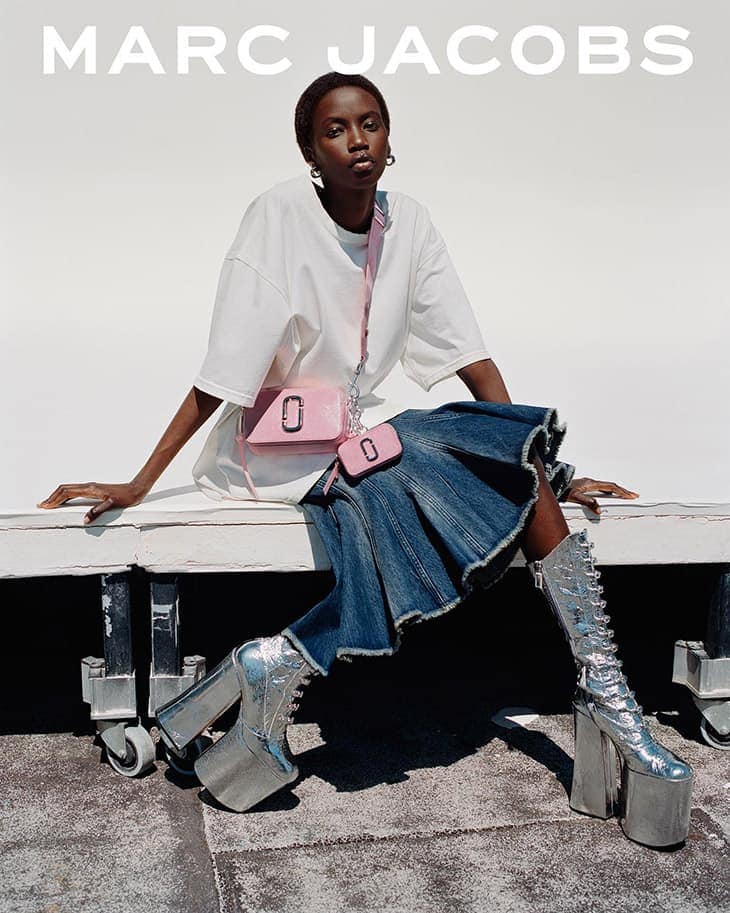 Fashion editorial shot of a Black female model wearing an oversized white T-shirt, blue denim skirt, and silver platform boots, posing indoors.