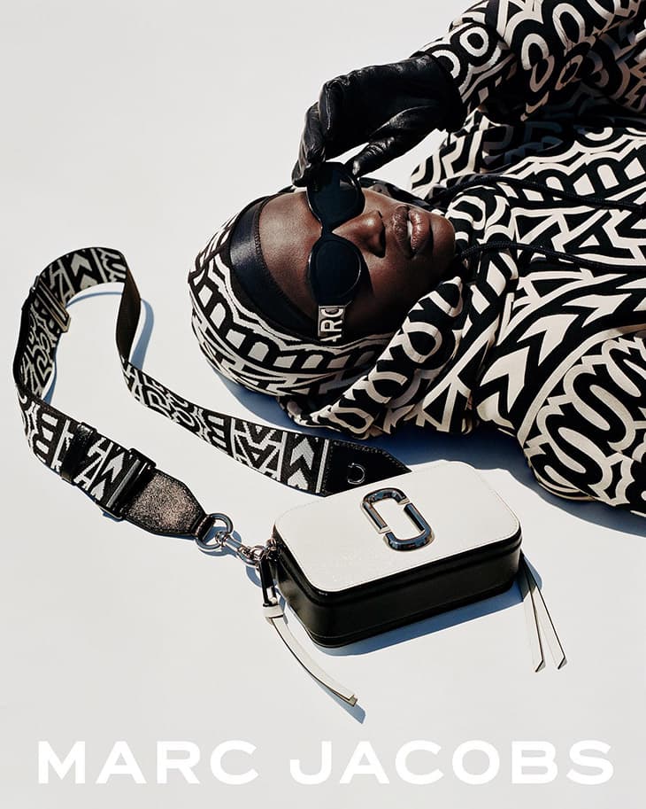 Fashion shot of an adult Black female model with sunglasses, headscarf, and a white Marc Jacobs handbag on a light backdrop.