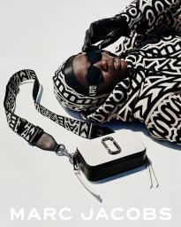 Anok Yai wearing patterned clothing and sunglasses with a white handbag against a plain background for Marc Jacobs