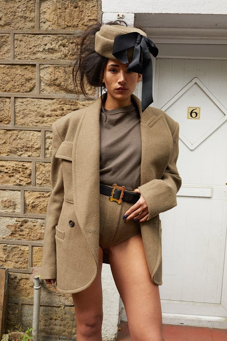 A woman in a neutral-tone outfit with a hat poses against a brick and white door backdrop, exuding confidence and vintage style.