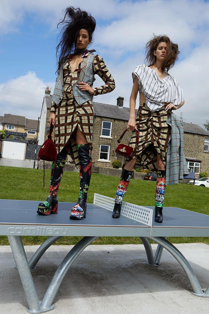 Two fashion models wearing dresses and platform boots stand on a ping pong table outdoors on a sunny day with a blue sky backdrop.