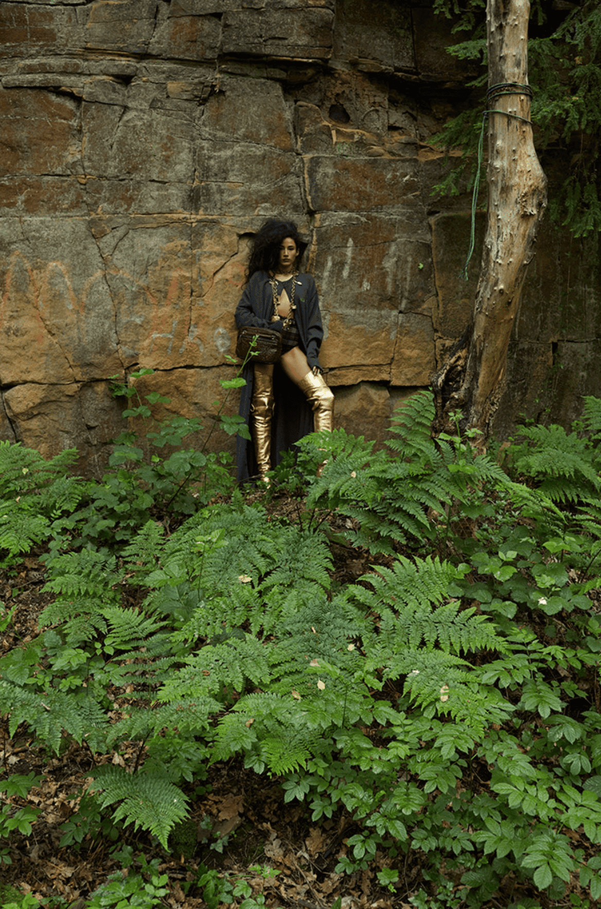Woman with afro poses in gold thigh-high boots and long coat near rock face surrounded by lush green ferns and a single tree.