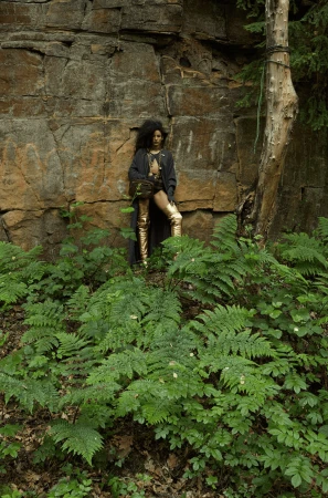 Woman with afro poses in gold thigh-high boots and long coat near rock face surrounded by lush green ferns and a single tree.