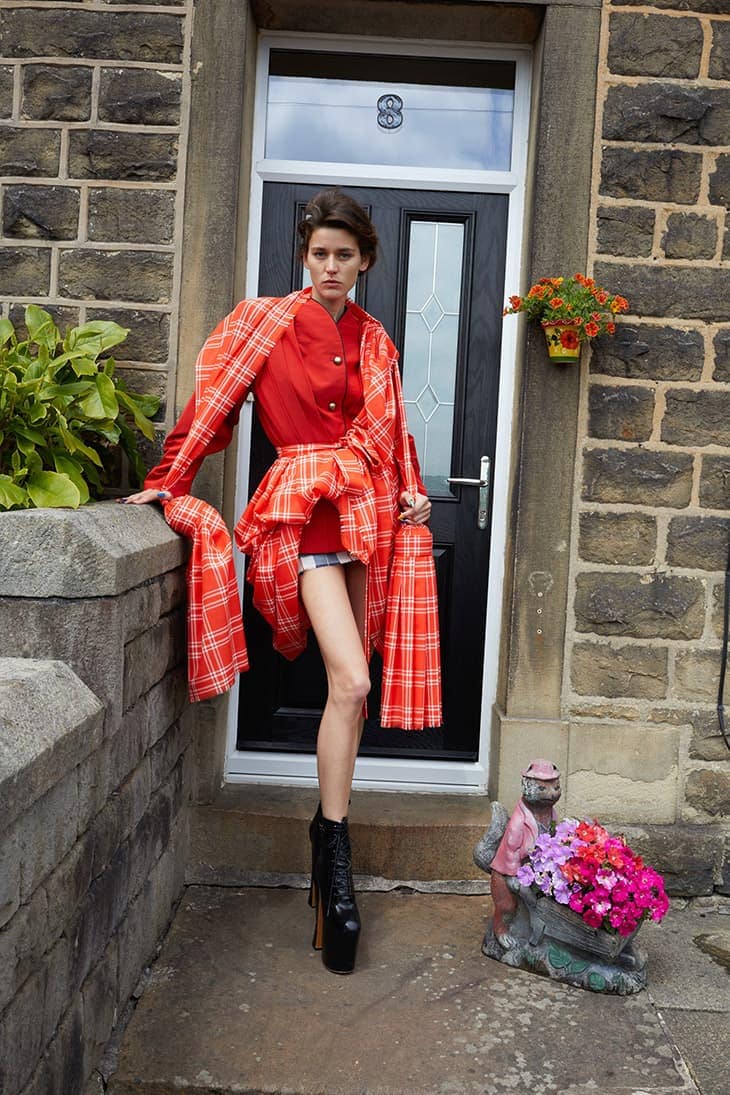 Fashionable woman in red outfit and platform boots poses on steps outside a stone house with a black door and potted flowers.