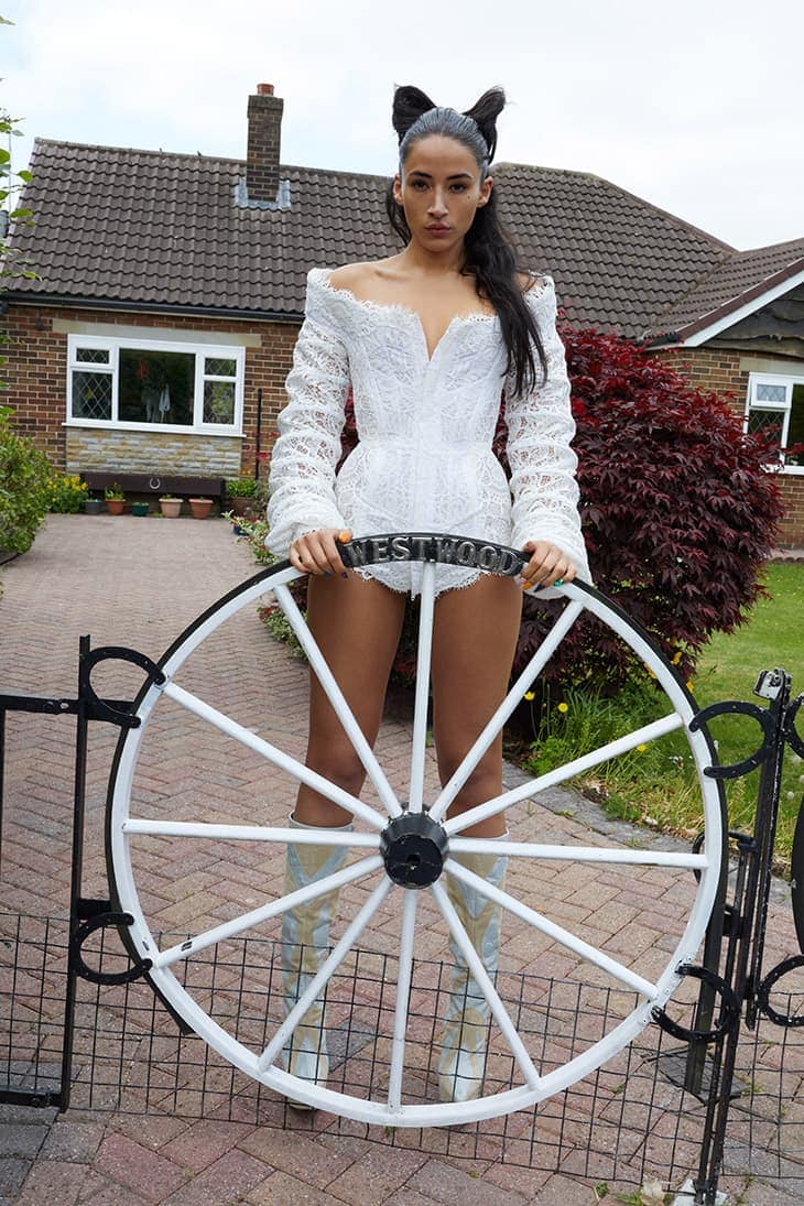 Woman in a white lace corset and boots standing in front of a house, holding a white wagon wheel displaying the word 'Westwood'.