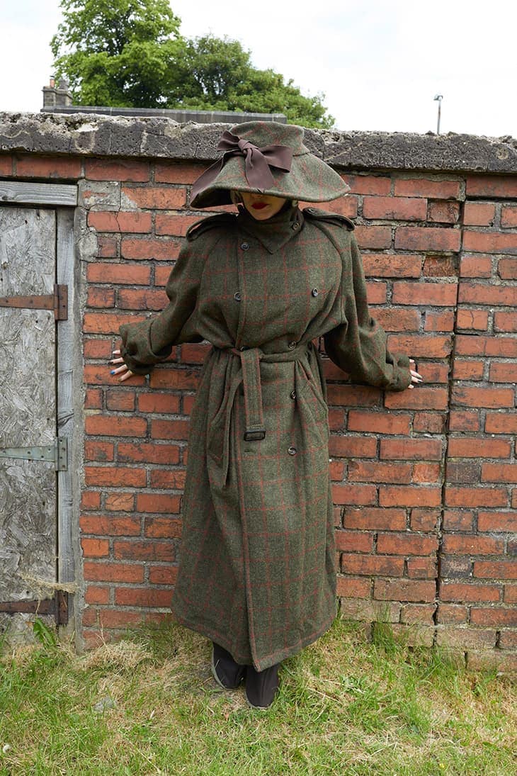 A person stands in front of a brick wall wearing a green coat, hat with a brown ribbon, and black shoes with outstretched arms.