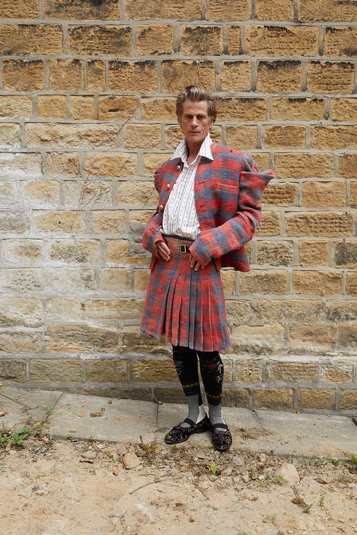 Full-body shot of an adult man in traditional Scottish attire: red tartan kilt and jacket, patterned stockings, and dark flats outdoors.