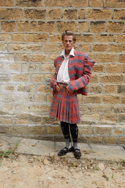 Full-body shot of an adult man in traditional Scottish attire: red tartan kilt and jacket, patterned stockings, and dark flats outdoors.