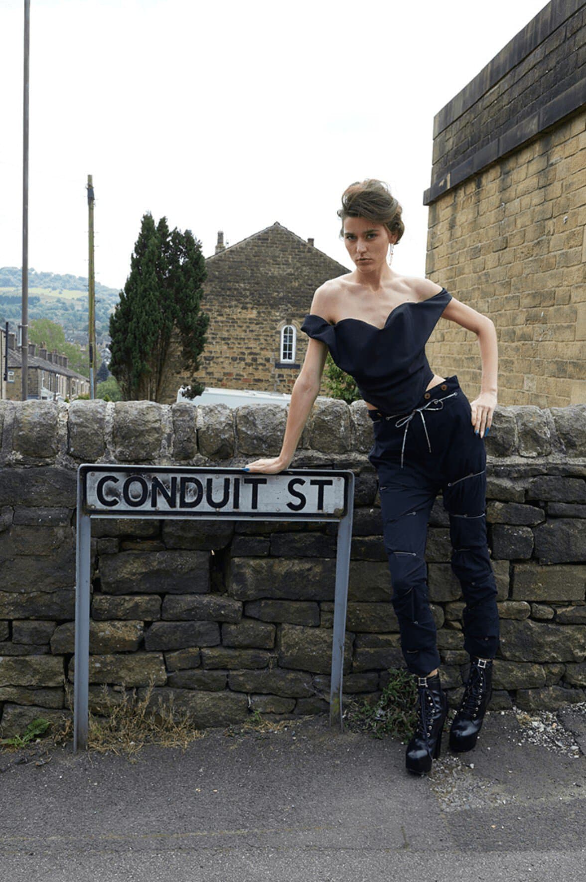 Fashion model poses leaning against street sign in an urban setting wearing a dark top, dark pants, and platform boots.