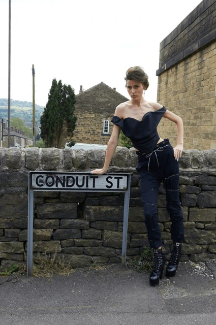 Fashion model poses leaning against street sign in an urban setting wearing a dark top, dark pants, and platform boots.