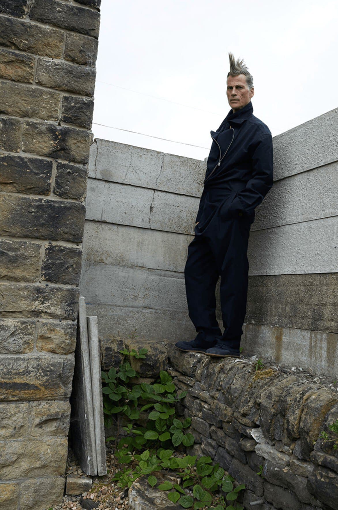Adult male with a mohawk, wearing a dark jumpsuit, stands on a low stone wall in an outdoor setting with a stone wall backdrop.