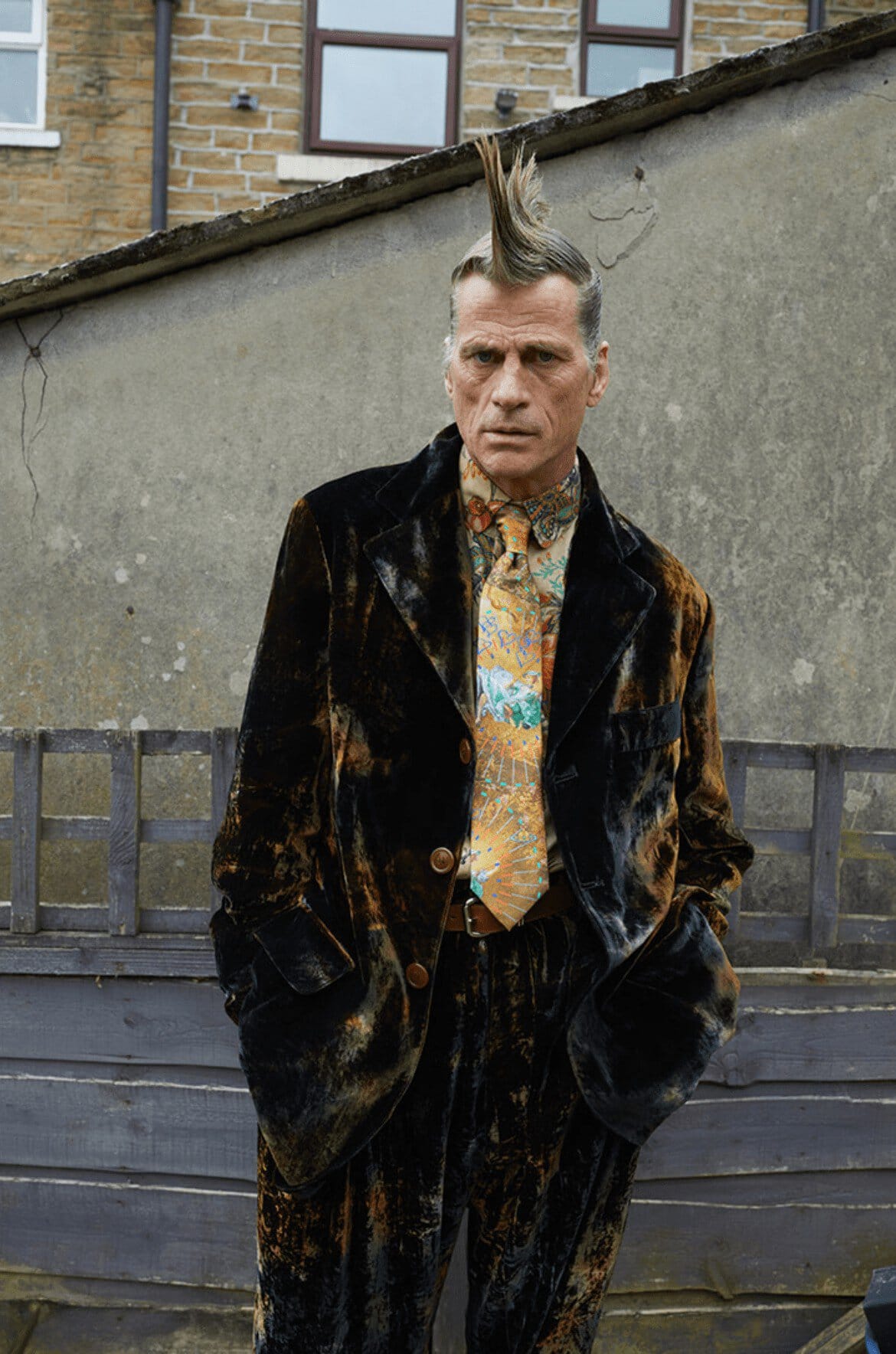 A man with a mohawk stands outdoors wearing a velvet suit and patterned tie, posing against a weathered wall and a glimpse of a building.